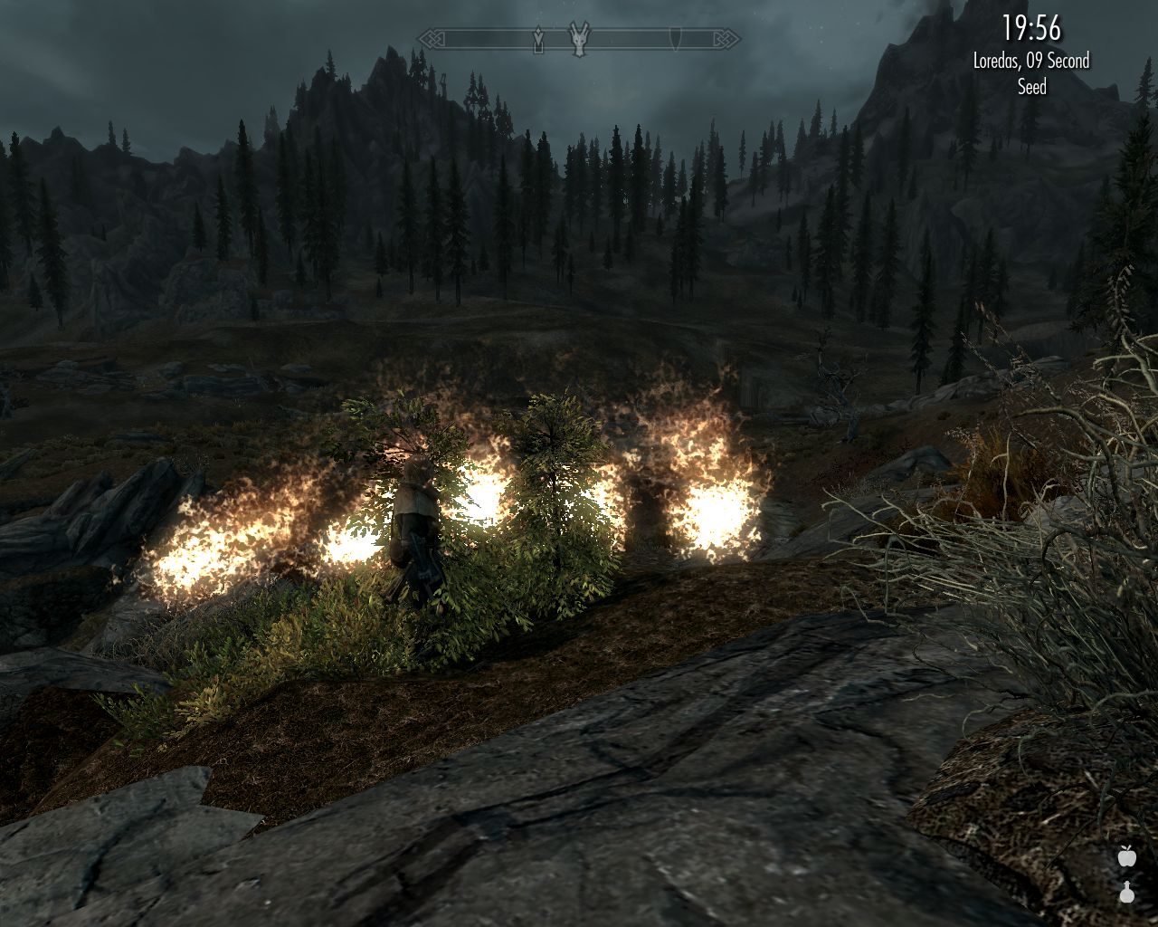 evening, downhill slope with rocks and grass, a man stands next to a large fire