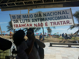 Foto com um casal dançando, um placa com a frase 18 de maio dia Nacional da Luta Anti-manicomial, trancar não é tratar. No fundo a praia de Iracema, em Fortaleza. 