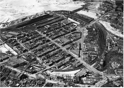 Black-and-white aerial photo of LeBreton Flats prior to it being expropriated. Wellington Street runs along the bottom of the photo from east to west. Booth Street crosses diagonally to the Chaudière crossing at the upper right. Four-lane Pooley's Bridge intersects with Wellington at the lower right.