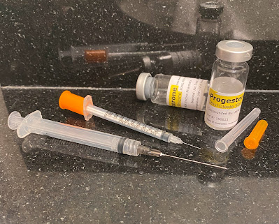 Photograph of two syringes (one larger than the other with a longer needle) and two bottles of progesterone (one empty and one half full) on a black bathroom counter