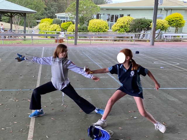 School playground with low trees in the background. Jemima, on the left, wearing long black pants, cyan sneakers, white fencing jacket and foil lamé demonstrates a fencing lunge. Her peer, on the right, dressed on school uniform, tries a lunge, holding a fencing foil. A fencing mask lies on the ground.