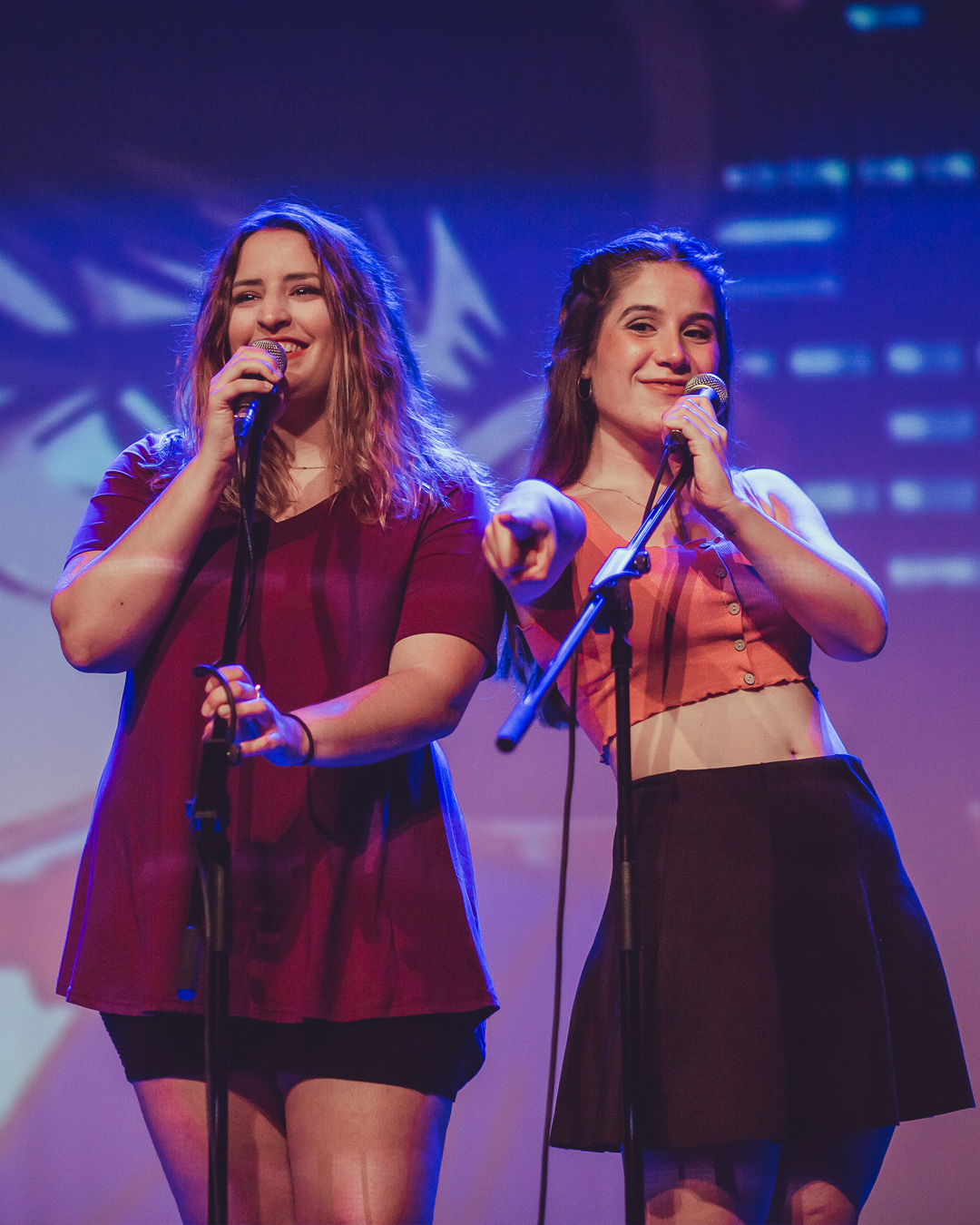 Dos chicas sonrientes con un micrófono en la mano señalan al publico