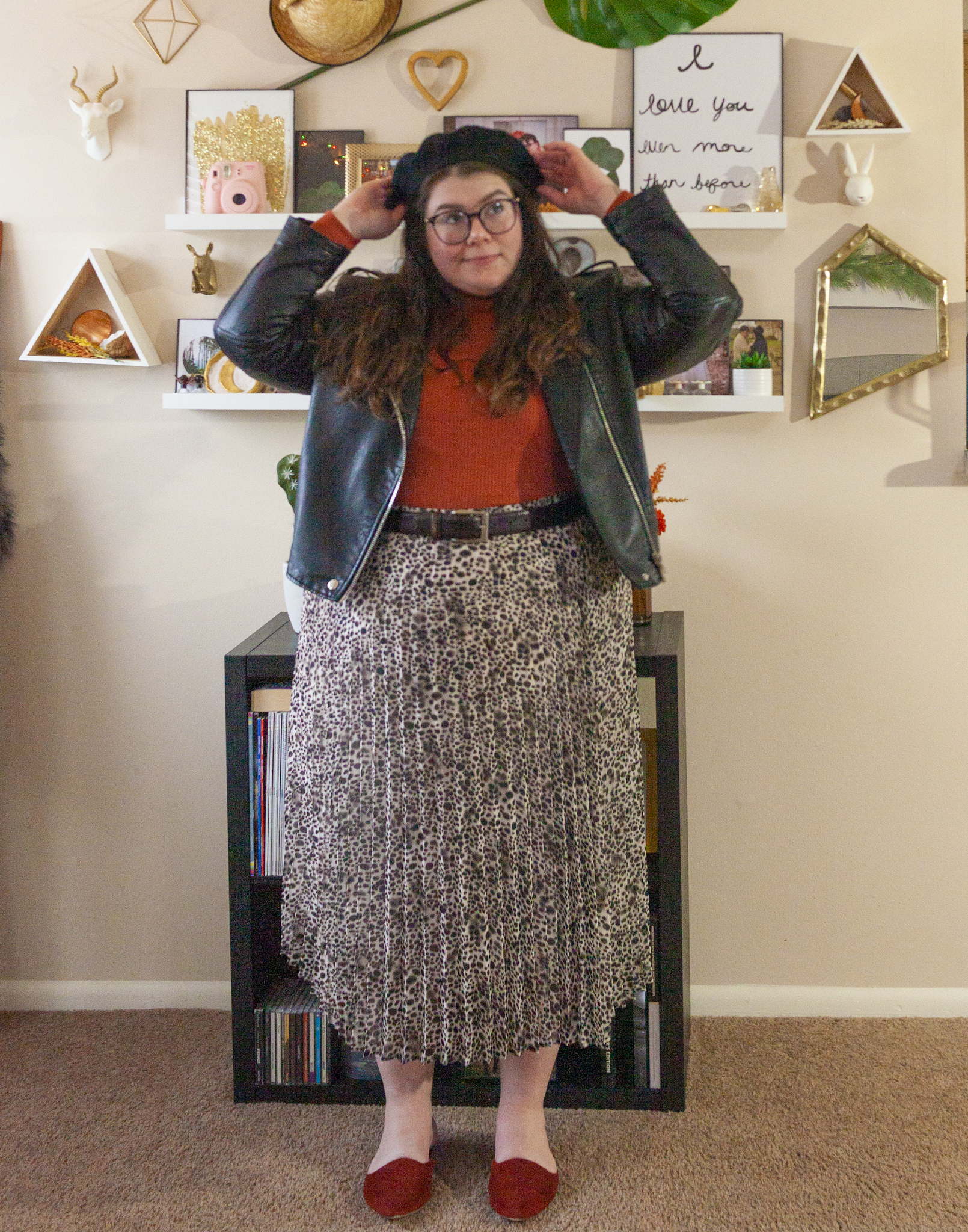 An outfit consisting of a black wool beret, a black faux leather moto jacket unzipped over a burnt orange mock neck long sleeve ribbed top tucked into a white and various shades of muted brown leopard print pleated midi skirt and burnt orange mule slides.