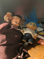 A short doll with plastic face wearing a velvet dress with white beads around her neck who is holding the most adorable blue-haired troll baby ever.