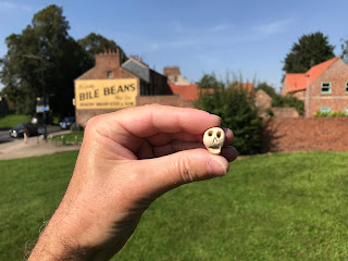 A picture of a hand holding a small ceramic skull with the Bile Beans advert on the side wall of the building in the background.  The ceramic skull being Skulferatu #49.  Photo by Kevin Nosferatu for the Skulferatu Project.