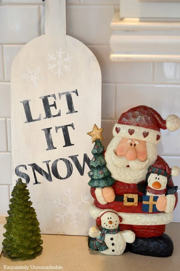 Let It Snow Wooden Sign sitting on countertop near Santa figurine and tree candle