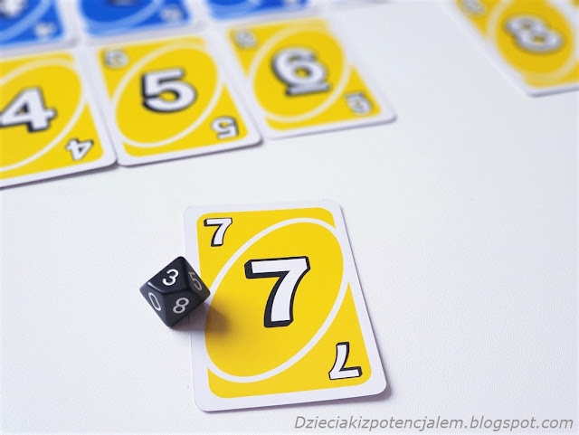 Na zbliżeniu widać kartę Uno w kolorze żółtym o wartości 7 oraz kostkę dziesięciościenną z wylosowaną wartością 3. obie wartości dają sumę 10 czyli cel gry