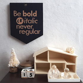 1/12 scale modern miniature tabletop scene in cream and brown containing a tiny bottlebrush Christmas tree, a lidded bowl with gold and white stripes, wooden blocks with the numbers '1' and '5' on them, and an empty dollshouse. Above it on the wall is a wallhanging that says 'Be bold or italic, never regular'