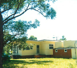 A tall tree frames the left hand side of the photo. Beyond is a side view of the front porch of the yellow building with red brick foundations. In the middle of the building is the back door. On the right hand side is a red brick tank stand with two cream-coloured water tanks on top and a hills hoist rotary washing line in the foreground.