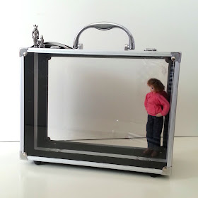 Beauty case with clear sides and a one-twelfth scale modern doll inside. On top of the case are two small metal ornaments.