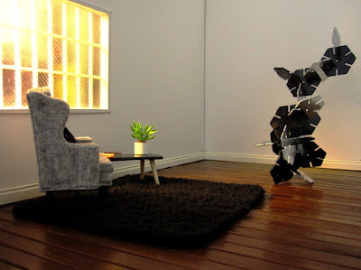 Modern miniature gallery space with a grey wing chair on a black rug facing a silver and black sculpture.