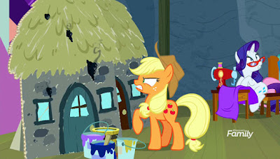 Applejack looks angrily at a fire-damaged prop house to the left, while to the right Rarity looks worried at something out of shot as she sits at her sewing machine