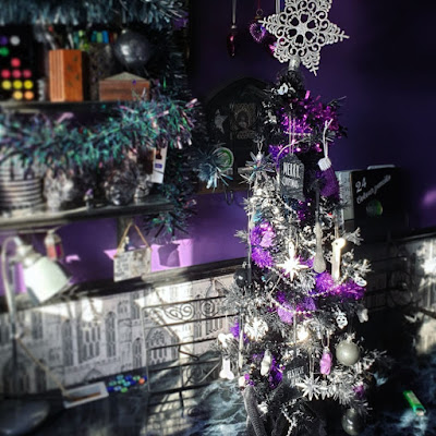 Christmas decorations in daylight A photograph of Christmas decorations. The wall behind is purple, there is a black dado rail, and greyscale wallpaper of Gothic arches. There are three shelves on the right holding art materials, and the upper two shelves are garlanded with tinsel, grey at the top, iridescent green and purple in the middle. There is a 2 and a half foot tall black Christmas tree densely decorated, with a glittery snowflake on the top that is too large and leans slightly to the right. The tree is on a desk with a fake black marble work-surface. The bottom of the tree is wrapped in a Hallowe'en table-cloth of black lace. The tree is decorated in a purple and silver colour-scheme, with skulls and stars as the primary motifs; it has lots of tinsel. Behind the tree is an ornate black metal stand for a tablet computer. There is a grey vintage-style desk-lamp on the desk.