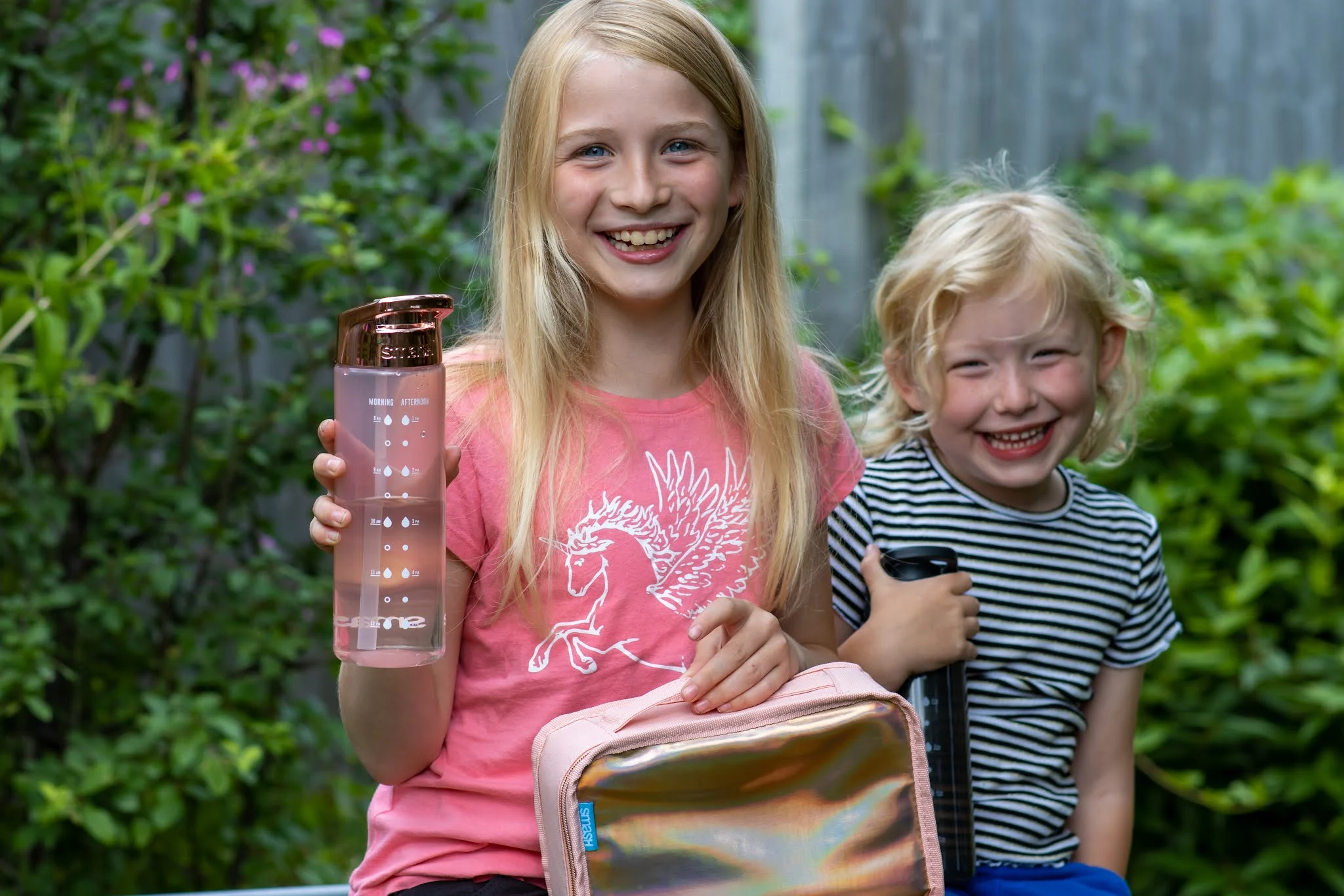 Two girls in a garden one with a SMASH rose gold water bottle and rose gold iridescent lunch bag, the other with a black water bottle
