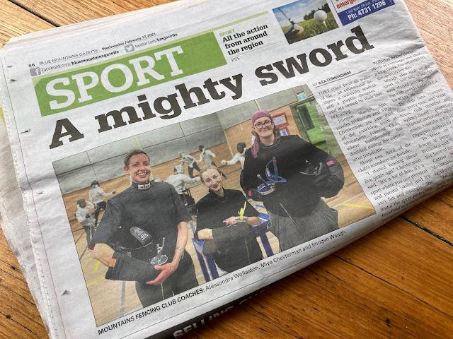 Newspaper folded to show the upper half of the back page, on which is a headline ‘A mighty sword’ and a photo of the three coaches facing the camera and smiling. In the background of the photo are several fencers are bouting. The easter egg is two of the members facing eachother and fighting with different weapons – left fencer with sabre and right fencer with epee. Obvs this is never done, so now you know that all the fencers in the background were called back as they were packing up after a busy training session to pose for this pic!