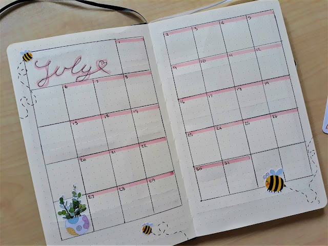 This page shows a calendar for July. There are boxes for each day with ping highlight over the date to make it brighter. July is written in calligraphy at the top left corner. There are bee stickers dotted around.