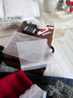 Modern dolls' house miniature bed with a suitcase and range of clothes and toiletries on it. On the suitcase are two zip lock bags.