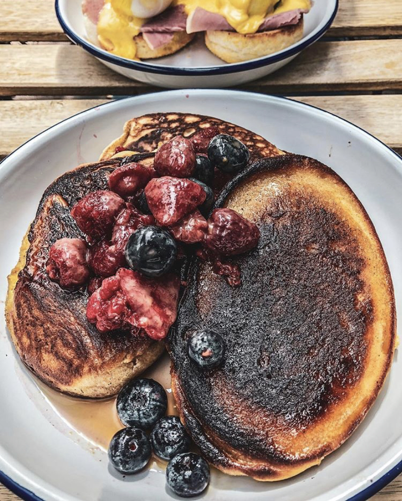 A stack of pancakes covered in maple syrup and summer berries on a white plate on top of a wooden table 