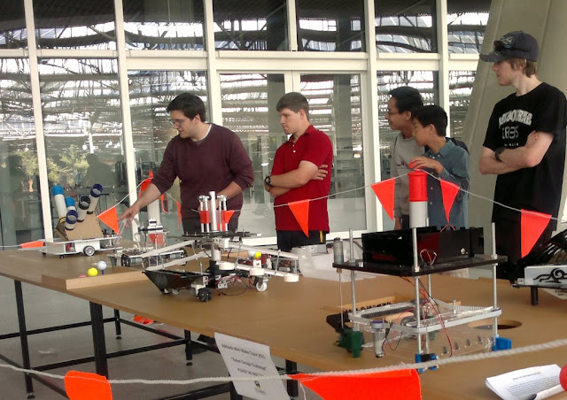 Four university students have their robots lined up on a table in front of them.