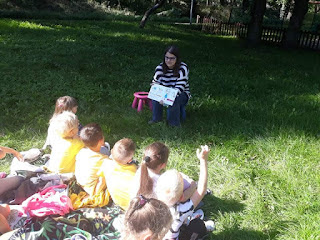 Pani pokazuje dzieciom siedzącym na kocu na trawie ilustracje w książce.