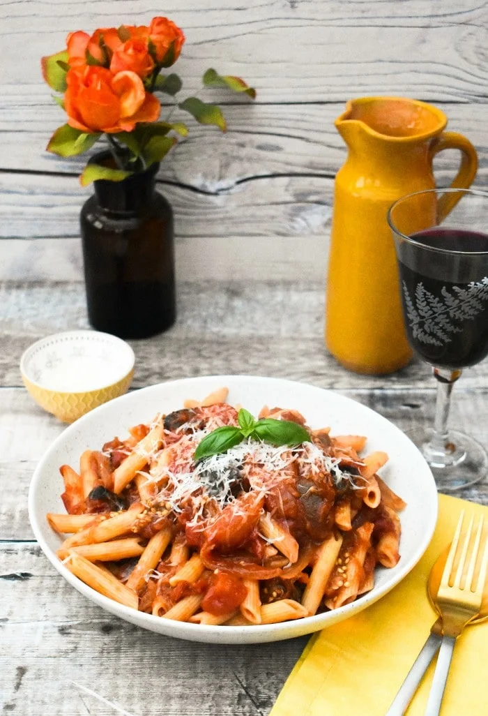 Aubergine tomato pasta in a pasta bowl on a table set with flowers, wine and a water jug
