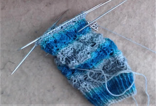 A textured sock on double-pointed needles.  The sock is striped blue/cream/grey.  A small celtic knot stitch marker is clipped into work. 