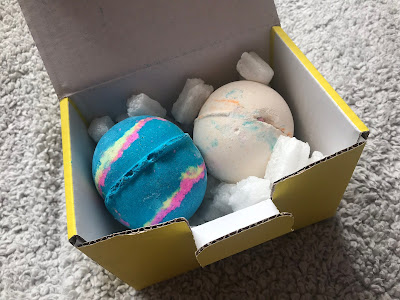 a white bath bomb and a blue bath bomb in a yellow cardboard box on a grey blanket
