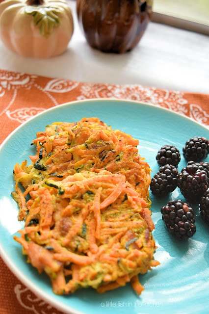 Two eye-catching sweet potato and zucchini cakes are served along side a portion of vibrant ripe blackberries an a teal ceramic plate. The sweet potato cakes are delightly colorful: shredded amber sweet potato is mingled with bits of green from the zucchini mixed in perfectly formed round, veggie pancakes.