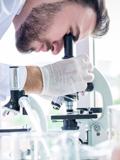 homem com cabelo penteado para trás e barba que tentou limpar em um laboratório científico com luvas de borracha ao olhar em um microscópio