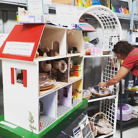 Child's wooden dolls' house filled with home-made furnishings, on display next to a shelving unti displaying baskets of craft items. A woman is looking through one of the baskets.