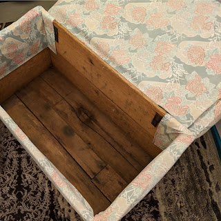 A photo taken from above, showing the inside of an ottoman with the lid open, it is made from treated timber inside and the fabric stapled around the edges. The fabric is less faded here, but still aged and very old fashioned.