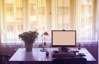 A desk with flowers, a lamp, and a computer in front of a window with sheer curtains