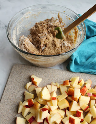 bowl of batter for apple cake behind cutting board full of chopped apples