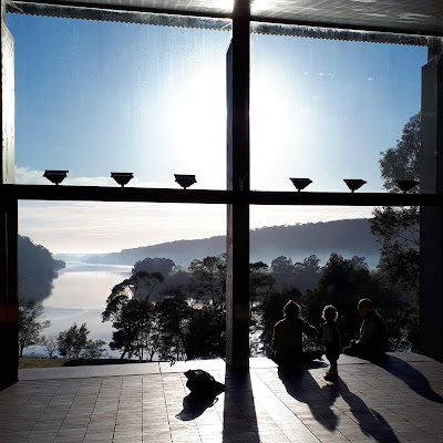 View from inside the main block of the Boyd Education Centre, looking through the open sidling doors down to the river. In the doorway are sillouheted two men and a child.