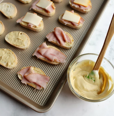 These are great warm, but are still really good at room temperature. They are a great food to set out for a party! baking sheet with baguette slices on it, some spread with mustard sauce, some with ribbons of ham on that and some with small slices of cheese on top building the crostini and getting them ready for the oven