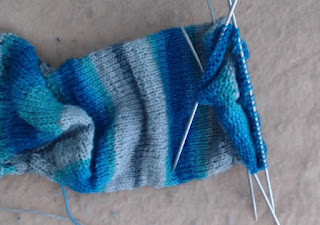 A knit sock on double-pointed needles.   The yarn is self-striping in shades of blues and greys. 