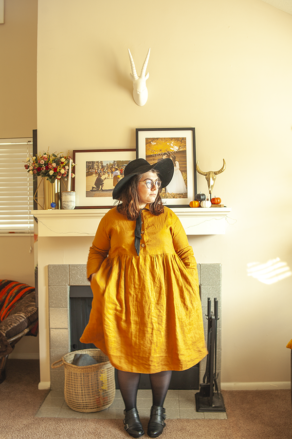 An outfit consisting of oversized black hat, yellow collared linen babydoll dress, sheer tights and black Chelsea boots.
