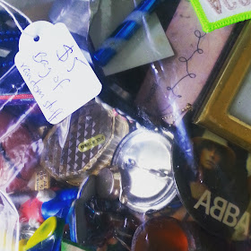 Plastic bag containing various small bits and bobs (including metal badges, small containers and frames) with a price tag saying '$5 Bag of randon stuff'' attached.