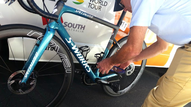 A metallic blue-green bike with white lettering ("Specialized" "S-Works") is leaning against a team bus.  The Technical Commissaire is leaning over it with a gadget in his hands that looks similar to a large mobile phone or iPad. The Technical Commissaire is wearing a dark blue hat, a light shirt and light brown trousers. The bus is white with sponsorship logos printed on it.The bike has a white tag attached to the seat post, underneath the seat with the number 11.