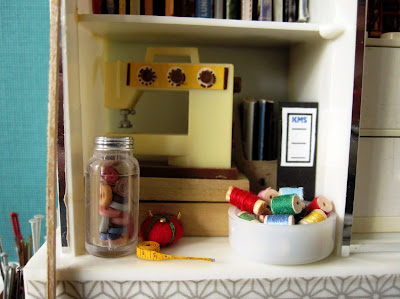 Details of a 1/12 scale miniature studio shelving unit containing a sewing machine, thread, buttons, a pin cushion and tape measure.