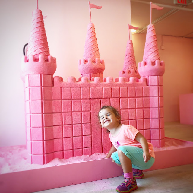 Toddler girl smiles in front of a pink sandcastle