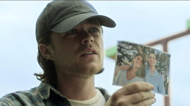 Guy with long hair wearing a hat and checkered shirt holding up an image of a lost man and woman