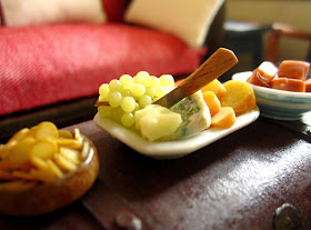 Miniature dolls' house finger food arranged on a coffee table, including a bowl of chips, a cheese and grape platter and a bowl of fudge,