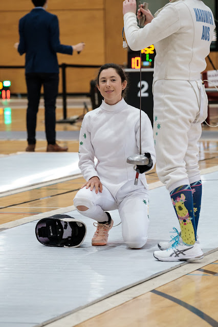 Marina Carrier kneels on the piste holding her weapon vertically as another fencer prepares to put the weight on the tip of her epee to check it. Marina looks calm and professional, well why wouldn't she, she's an an amazing athelete who is about to take home a medal!