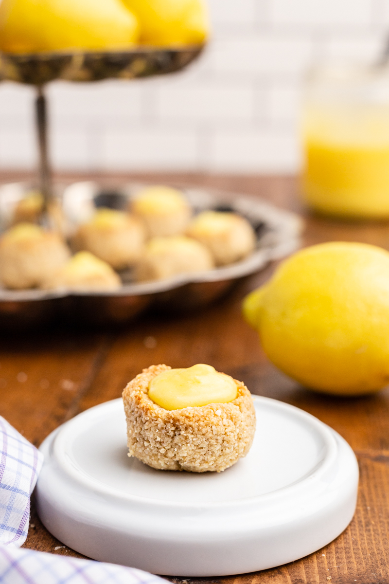 Photo of a Keto Lemon Thumbprint Cookies on a small white plate with a cookie stand full of them in the background.