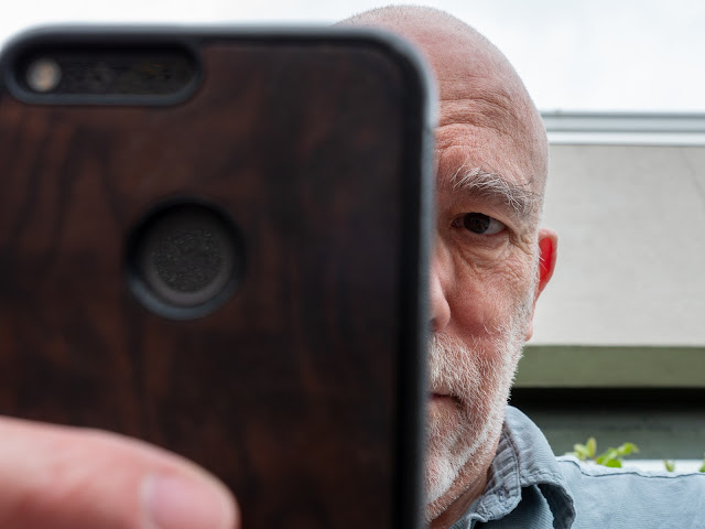 photograph of an older white male looking at the camera with his cell phone blocking half of his face