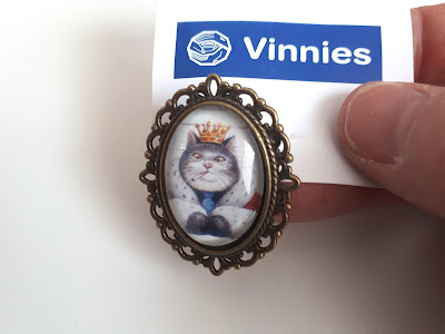 Vintage-looking portrait brooch with a drawing of a cat in a crown and robe, smoking a cigarette.