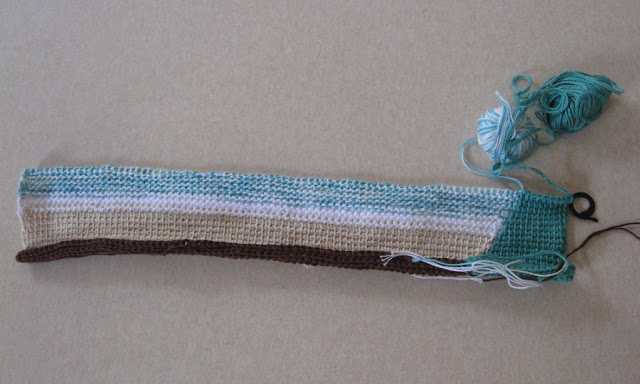 A placemat in progress worked from bottom to top. Stripes from the bottom up: brown, beige, white and variegated white/aqua. The right hand ends of each stripe are all worked in green.  The two current balls of cotton in variegated white/aqua and green are at top right. A black stitch marker on the top right hand edge of the mat keeps the work from unravelling.