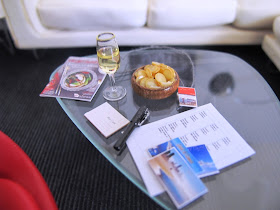 Modern one-twelfth scale miniature noguchi coffee table with a selection of postcards, a glass of wine and a travel magazine on it.