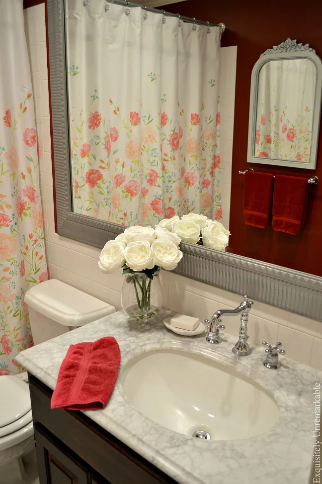 Red bathroom with floral shower curtain and flowers on vanity top with red towel draped over side.